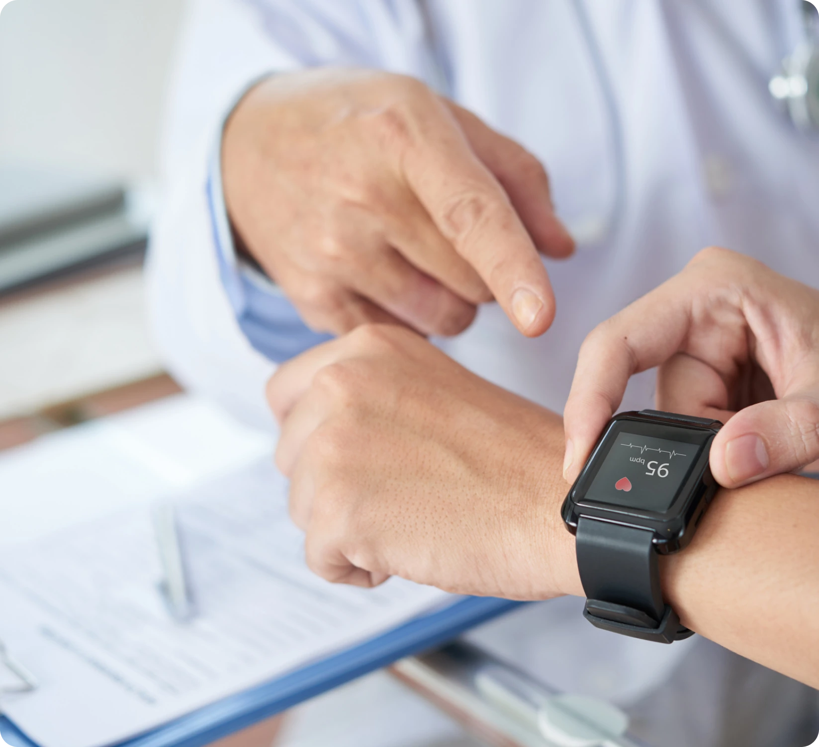 A doctor is pointing at a smartwatch displaying a heart rate of 95 on a person's wrist. A clipboard with papers is on the table.