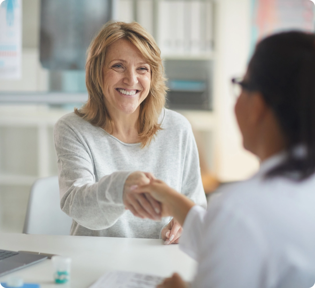 A woman smiles and shakes hands with a person in a medical coat in an office setting.