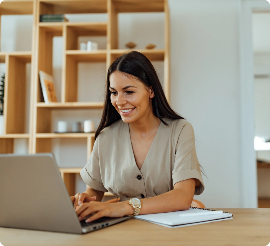 A woman with long dark hair is smiling while typing on a laptop at a desk. Notebooks are open beside her. A wooden shelf with books and decor is in the background.